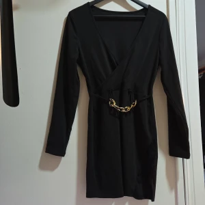 Festklänning Kort Svart Omlott Kedjedetalj, M - Säljer en figurnära och snygg kort klänning i klassisk omlottmodell (wrap-stil) framtill. Perfekt för fest, middag, utekväll eller när du vill känna dig extra uppklädd. ​Design: Omlott V-ringning och långa ärmar. Spets i kanten nertill. Klänningen har en insydd eller fastsydd midjedetalj med en iögonfallande guldfärgad kedja som lyfter designen.