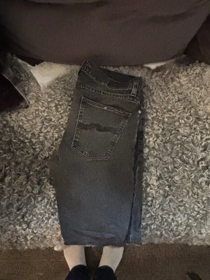 Svarta jeans från Nudie Jeans - Snygga svarta jeans från Nudie Jeans med slitna detaljer och klassisk femficksdesign. Jeansen har en rak passform, tydliga sömmar och Nudie-loggan på bakfickan. Materialet är robust denim och de har dragkedja framtill. Perfekta för vinter ❄️Ny pris: 1500kr. Mitt pris: 599kr.