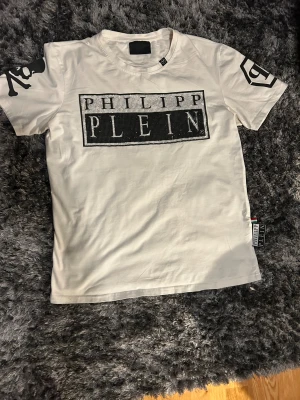 Vit Philipp Plein t-shirt med paljetter - Snygg vit t-shirt från Philipp Plein med stort svart och silvrigt paljettmärke på bröstet. Coola tryck på båda ärmarna, bland annat dödskalle och PP-logga. Gjord i mjuk bomull, klassisk passform och rund hals. Perfekt för dig som gillar statement-plagg.