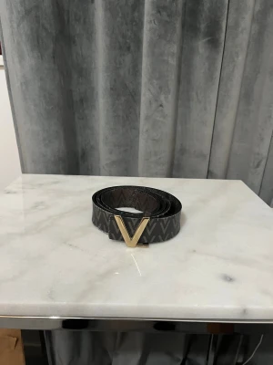 Svart skinnbälte med V-spänne - Snyggt svart bälte från Valentino med ett stort V-format spänne i guld. Bältet har ett mönster med gråa V-symboler längs hela remmen och är tillverkat i skinn. Perfekt accessoar för att lyfta din outfit med en exklusiv touch.