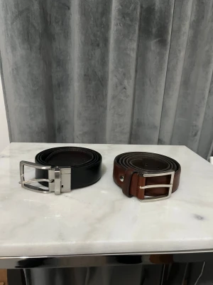 Svart och brunt skärp i skinn - Två stilrena bälten i svart och brunt skinn med klassiska silverfärgade metallspännen. Perfekt för att ge outfiten en clean och snygg touch. Båda har en enkel, rak form och passar till jeans eller kostymbyxor.