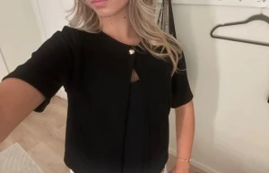 Svart topp med guldfärgad knapp - Snygg svart topp från Gina med korta ärmar och guld detalj. Passar perfekt nu till jul och andra roligheter. 