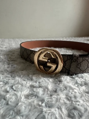  bälte med guldspänne - Snyggt bälte med klassiskt mönster i beige och brunt. Bältet har ett stort, rundat spänne i guld med dubbla G-loggan. Perfekt accessoar för att lyfta vilken outfit som helst.