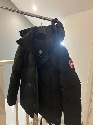 Svart dunjacka från Canada Goose - Svart dunjacka från Canada Goose med klassisk patch på ärmen, stor huva och rejäla fickor. Jackan har dragkedja och knappar framtill, samt breda quiltade sektioner som ger en puffig look. Perfekt för kalla vinterdagar och riktigt snygg streetstil.