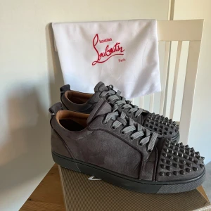 Christian Louboutin grå sneakers - ÄKTA Snygga grå sneakers från Christian Louboutin i mocka med ikoniska röda sulor och coola nitar på tån. Skorna har rund tå, platt sula och grå snörning. Perfekta för dig som vill sticka ut med exklusiv streetstyle. Tre  nitar saknas, Kvitto finns