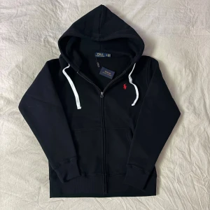 Svart hoodie från Polo Ralph Lauren - Svart hoodie från Polo Ralph Lauren med vit snörning och röd broderad logga på bröstet. Tröjan har huva, dragkedja framtill och långa ärmar. Tillverkad i mjuk bomull för skön komfort. Perfekt för chill dagar och streetwear vibes. Storlek S.