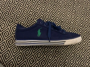Marinblå sneakers från Polo Ralph Lauren - Snygga marinblå sneakers från Polo Ralph Lauren med grönt broderat logomärke på sidan och 'POLO' tryckt bak. Skorna har snörning, rund tå och platt sula med vita och blå ränder. Ovandelen är i textil och insidan har gröna detaljer.