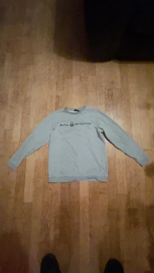 Grå Sail Racing sweatshirt - Snygg grå sweatshirt från Sail Racing med tryckt logga över bröstet. Tröjan har rund hals, långa ärmar och ribbade muddar vid ärmslut och nederkant. Perfekt för dig som gillar stilren och sportig design.