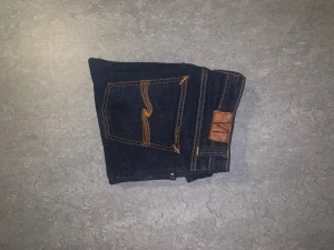 Mörkblå Nudie Jeans - Snygga mörkblå jeans från Nudie Jeans Co med orange söm och broderad bakficka. Modellen har rak passform med 31 W och 30 L, tillverkade i slitstarkt bomullsdenim.