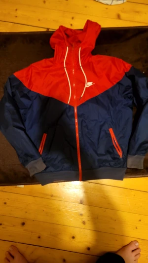 Nike vindjacka röd och marinblå S - Nike vindjacka i rött och marinblått med huva och vita snören. Jackan har dragkedja framtill, två fickor med röda dragkedjor och ribbade muddar vid ärmslut och nederkant. Perfekt för blåsiga dagar och sportig stil.
