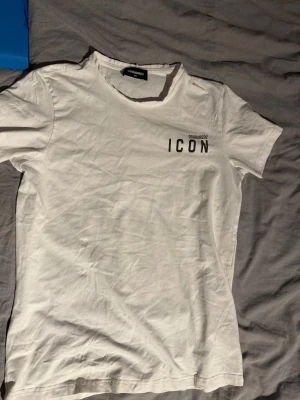 Vit ICON t-shirt från Dsquared2 - STORLEK 14Y! Snygg vit t-shirt från Dsquared2 med trycket 'ICON' i svart på bröstet. Klassisk rund hals och korta ärmar. Perfekt för dig som gillar stilrena och trendiga plagg med statement-tryck. Tillverkad i mjuk bomull för skön känsla hela dagen.