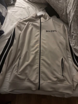 Beige track jacket från Palm Angels - Säljer en beige track jacket från Palm Angels med svarta ränder längs ärmarna och logga på bröstet. Jackan har dragkedja framtill, två fickor med dragkedja och ribbade muddar. Perfekt för en clean och sportig streetwear-look.