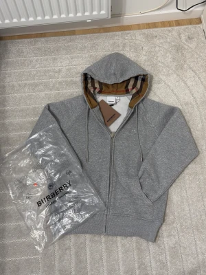 Grå Burberry hoodie med dragkedja - Grå hoodie från Burberry i storlek M med klassiskt rutigt foder i huvan. Tröjan har dragkedja framtill, snörning vid huvan och broderad Burberry-logga på bröstet. Mjuk bomullskvalitet och ribbade muddar. Perfekt för dig som gillar stilren design med lyxig touch. Storlek M men sitter som S och xs om man vill ha tajt