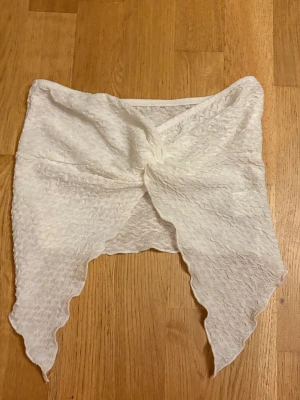 Vit bandeau tubtopp med knyt - Säljer en vit bandeau tubtopp med snygg knytning framtill och vågiga spetsiga kanter. Toppen har ett lätt strukturerat tyg som ger cool textur och är perfekt för sommarens varma dagar. Passar dig som gillar att sticka ut med detaljer och unik form.