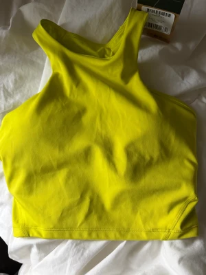 Gul träningslinnetopp från The North Face - Supersnygg gul linnetopp från The North Face i modellen Refina Longline Bra. Toppens färg är sulphur spring green och den har en sportig, croppad passform med racerback och tryckt logga på ryggen. Materialet är stretchigt och består av polyester med Lycra för extra komfort.