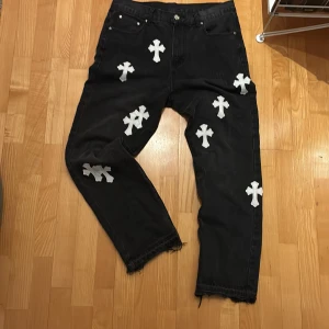 Levi's 501 x Chrome Hearts jeans  - Svarta Levi's 501 jeans med Chrome Hearts vita läderpatches i korsform över hela byxan. Klassisk femficksmodell med råa, fransiga benslut och silverdetalj på bakfickan. Snyggt straight fit och unik design för dig som vill sticka ut.