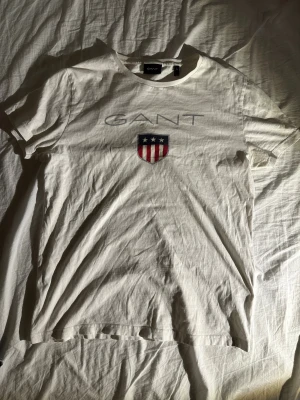 Vit GANT t-shirt med tryck, storlek S - Vit t-shirt från GANT i mjuk bomull, med klassiskt rund hals och korta ärmar. På bröstet finns ett stort GANT-tryck och ett sköldmotiv med stjärnor och röda ränder. Enkel och stilren design som funkar till jeans eller shorts.