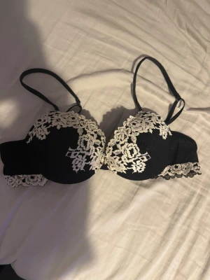 Svart spets-bh med blommig brodyr - troligen 75B💗 - Snygg svart bh med vita blommiga spetsdetaljer och brodyr över kuporna. Axelbanden är justerbara och kanten har extra spets för en fin touch. Perfekt för dig som gillar en feminin och elegant stil.