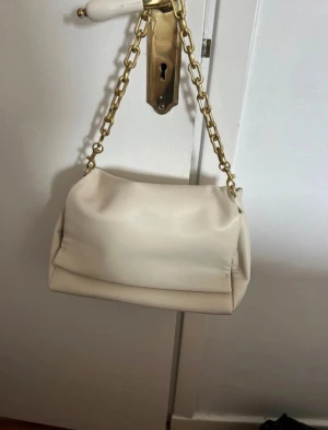Crémevit axelväska med kedja från Zara - Axelväska från Zara med mjuk, slät yta i skinnimitation. Väskan har en chunky guldfärgad kedja som axelrem 