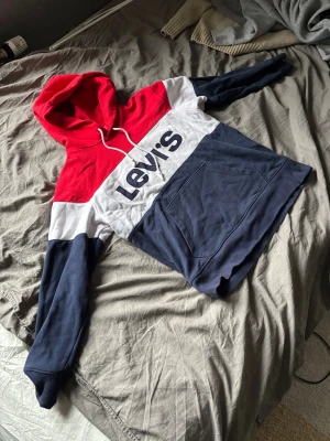 Levi's hoodie i rött, vitt och blått - Snygg hoodie från Levi's i storlek M med breda färgblock i rött, vitt och mörkblått. Stor logga framtill, huva med snörning och magficka. Mjukt bomullsmaterial och långa ärmar. Perfekt för chill dagar och streetstyle.