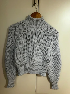 Ljusblå stickad polotröja - Chunky stickad polotröja i ljusblå med breda ribbade muddar och hög krage. Tröjan har en loose passform och grovstickad struktur som ger en cool och mysig vibe. Perfekt att slänga på sig när det är kallt ute.