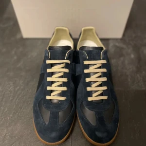 Maison margiela Gats - Snygga mörkblå sneakers med beige snörning och gummisula. Ovandelen är en mix av mocka och skinn, vilket ger en schysst kontrast. Insidan är ljus och sulan har ett coolt mönster undertill. Perfekt för dig som gillar stilrena och klassiska sneakers. Kvitto och dustbag medföljs! 