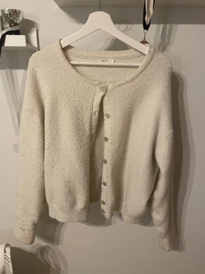 Vit kofta från MILOS med knappar - Mysig vit kofta från MILOS med rund hals och knäppning framtill. Koftan har en fluffig, stickad känsla och långa ärmar. Perfekt för att hålla sig varm och ändå se chill ut. Passar dig som gillar enkel och clean stil.