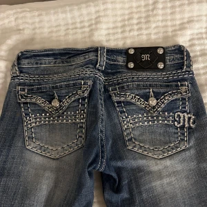 Miss Me blå bootcut jeans med detaljer - Snygga blå jeans från Miss Me med bootcut passform och låg midja. Jeansen har coola broderade fickor bak, dekorerade med pärlor och bling samt en metallplatta med logga. Materialet är klassisk jeans med slitningar och kontrastsömmar för extra edge.