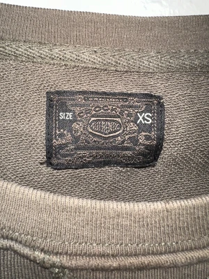 Olivgrön sweatshirt Iskenderian XS - Cool olivgrön sweatshirt från Iskenderian i storlek XS. Tröjan har ett stort tryck med bilmotiv och text framtill, rund hals och långa ärmar. Perfekt för dig som gillar retrovibbar och streetstyle. Materialet känns mjukt och skönt mot huden.