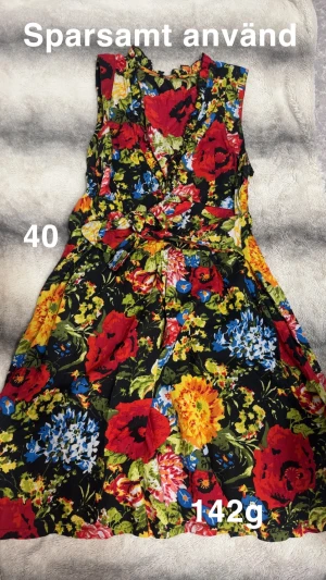 Blommig klänning med knyt - Färgstark klänning med knyt i midjan och blommigt mönster i rött, blått, gult och grönt. Ärmlös design och lätt, flödande tyg som ger en skön känsla. Perfekt för dig som gillar att sticka ut med färg och mönster.🌸 Sparsamt använd. Storlek 40