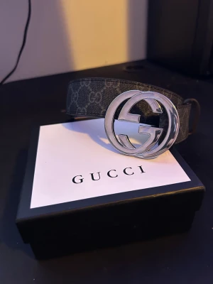 Gucci GG Supreme bälte med silverfärgat spänne - Säljer ett Gucci GG Supreme bälte med ikoniskt silverfärgat GG-spänne. Bältet har svart och grått monogrammönster på utsidan och ljusbrun läderinsida. Kommer med originalbox, kvitto och kontrollkort. Perfekt för dig som vill ha en lyxig detalj till outfiten.