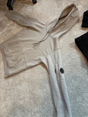 Beige hoodie från C.P. Company - Snygg beige hoodie från C.P. Company med klassisk huva och snörning. Tröjan har en stor magficka och en cool detalj med glaslins på ena ärmen. Tillverkad i mjuk bomull, perfekt för chill dagar. Passformen är normal och ärmarna är långa.