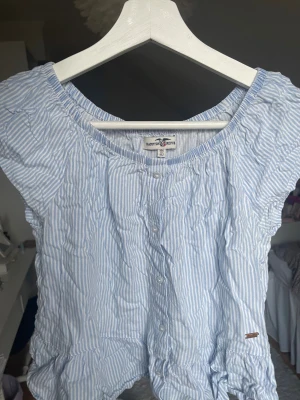 Blårandig blus från Hampton Republic - Säljer en söt blus från Hampton Republic i storlek 146/152. Blusen är ljusblå och vit med smala ränder, har korta ärmar och knappar framtill. Rundad hals och volangdetaljer längst  nere. Om ni vill ha bättre bilder med tanke på att blusen är lite skrynklig är det bara att säga till! 💙🤍Pris går att diskutera!
