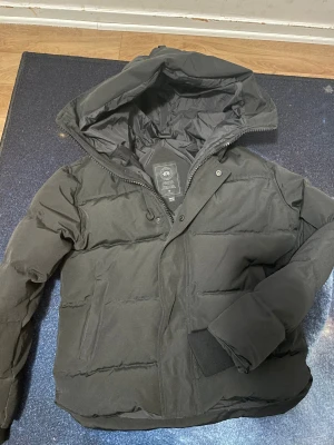  Canada Goose MacMillian  - Säljer en svart pufferjacka från Canada Goose i storlek M. Jackan har huva, ribbade muddar och dragkedja med tryckknappar. Klassisk Canada Goose-märke på ärmen och quiltad design. Perfekt för kalla dagar och riktigt snygg stil.