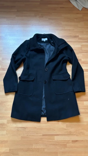 Svart kappa från Amica med ribbad krage - Svart kappa från Amica med snygg ribbad krage och klassisk knäppning framtill. Jackan har två stora fickor med lock och är helfodrad med slätt tyg på insidan. Långärmad och med stilren, rak passform som ger en clean look. 