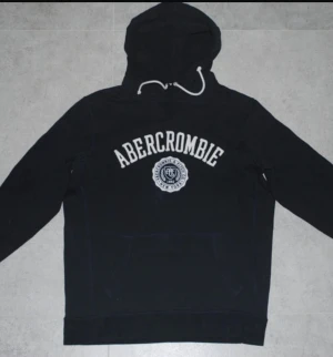 Blå hoodie Abercrombie & Fitch M - Mörkblå hoodie från Abercrombie & Fitch i storlek M. Tröjan har vit broderad logga och emblem på bröstet, huva med vit snörning och en klassisk känguruficka framtill. Perfekt för chill dagar och streetwear-stil.
