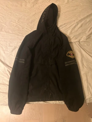 Svart vindjacka från Zavetti Canada - Svart vindjacka från Zavetti Canada med huva och dragkedja framtill. Jackan har ett snyggt patch med lönnlöv på insidan och logga på ärmen. Reflekterande ränder på båda ärmarna och elastiska muddar. Materialet är syntet och jackan har snörning nedtill. Det är dock en liten fläck på magen neråt men inget stor fläck ( jag kan sänka priset vid snabb affär)