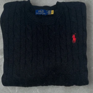 Svart kabelstickad tröja Polo Ralph Lauren - Svart kabelstickad tröja från Polo Ralph Lauren i storlek M. Klassisk rund hals och långärmad modell med röd broderad logga på bröstet. Tröjan är gjord i mjuk bomull och har ribbade muddar vid ärmslut och nederkant. KVITTO FINNS 