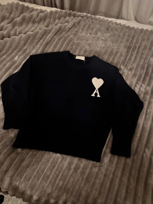 Svart stickad tröja från Ami Paris - Mörkblå stickad tröja från Ami Paris med vit hjärtformad A-logga på bröstet. Rund halsringning och ribbade muddar vid ärmslut och nederkant. Klassisk och stilren design som är enkel att matcha💯💯