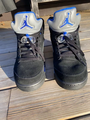 Nike Air Jordan 5 Retro svart/blå - Nike Air Jordan 5 Retro sneakers i svart mocka med blå och silver detaljer. Höga sneakers med snörning, genomskinlig lace lock och ikonisk Jumpman-logga på plösen och hälen. Sulan har blå och grå inslag, och meshpaneler på sidorna ger extra attityd.
