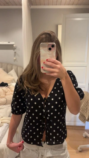 Svart prickig blus från Dobber - Svart blus med vita prickar från Dobber i storlek XS. Blusen har lite puffiga korta ärmar, v-ringad hals och snörning framtill. Materialet är lätt och luftigt. Passformen är normal och blusen är jätte fint croppad.