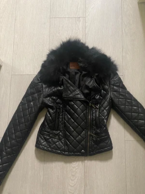 Svart quiltad skinnjacka med päls - Snygg svart quiltad skinnjacka som är figursydd från adL med fluffig päls vid kragen. Jackan har flera dragkedjor, långärmade ärmar och en croppad passform. Materialet är syntetiskt skinn och päls, med coola detaljer och ett stilrent rutmönster. För dig som vill att det ska sitta snatched och har storlek 36 så kommer denna passa perfekt, men såklart också för de som bär på 34.