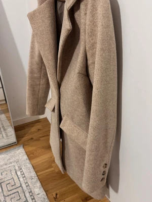 Beige kappa från Amisu i ullmix - Snygg beige kappa från Amisu med klassisk krage och knappstängning framtill. Jackan har två stora fickor med lock och stilrena detaljer vid ärmslutet. Perfekt för dig som gillar en clean och tidlös look under höst och vinter.