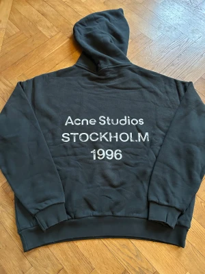 Svart hoodie Acne Studios Stockholm - Svart hoodie från Acne Studios med stor vit text 'STOCKHOLM 1996' på ryggen. Perfekt för chill dagar. Snygg och enkel design med street vibe. Skriv om frågor eller prisförslag!
