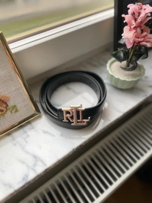 Svart skärp från Ralph Lauren - Stilrent svart bälte från Ralph Lauren i skinn med en bred, slät form och ett stort RL-spänne i guld. Bältet har klassisk design och justerbara hål för perfekt passform. Perfekt accessoar för att lyfta din outfit med en lyxig touch.