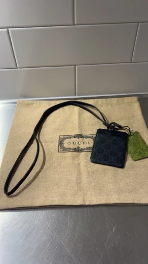 Svart plånbok med axelrem från Gucci - Snygg svart plånbok från Gucci med axelrem, perfekt för att bära runt halsen eller över axeln. Plånboken har GG-mönster och är tillverkad i skinn. Kommer med original dustbag och tag. Stilren och praktisk accessoar för dig som gillar designer.