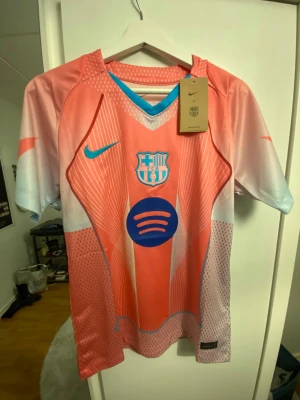 Barcelona Nike fotbollströja rosa/blå - Säljer en Barcelona fotbollströja från Nike i en snygg rosa och ljusblå färg med vita detaljer. Tröjan har korta ärmar, V-ringad krage med blå kant, och coolt grafiskt mönster framtill. Materialet är lätt och andas, perfekt för träning eller match.