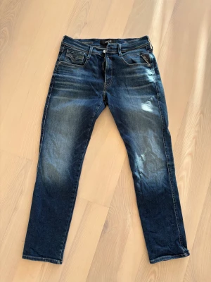 Replay Anbass jeans blå tapered W31 L30 - Snygga Replay Anbass jeans i mörkblå tvätt med coola slitningar och kontrastsömmar. Modellen är tapered med normal passform och klassiska fem fickor. Materialet är stretchigt jeans och de har dragkedja i gylfen. Perfekta för dig som gillar stilrena detaljer.