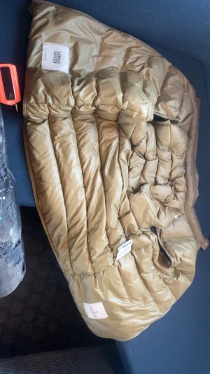 Beige dunväst från Moncler - Säljer en beige dunväst från Moncler med quiltad design och glansig finish. Säljer den för att dragkedjan är sönder går inte och stänga. Västen har en stilren look och är perfekt för lager-på-lager. Märkt med 'LONGUE SAISON' och har klassiska Moncler-taggar. Passar dig som gillar exklusiva och snygga ytterplagg.