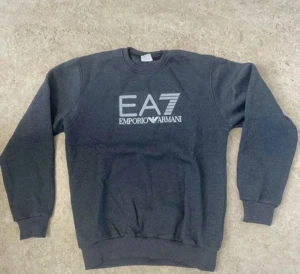 Mörkgrå EA7 Emporio Armani sweatshirt - Snygg mörkgrå sweatshirt från EA7 Emporio Armani med stort tryck på bröstet. Tröjan har rund hals, långa ärmar och ribbade muddar vid ärmslut och nederkant. Perfekt för dig som gillar stilrena och bekväma plagg med designer-vibe.